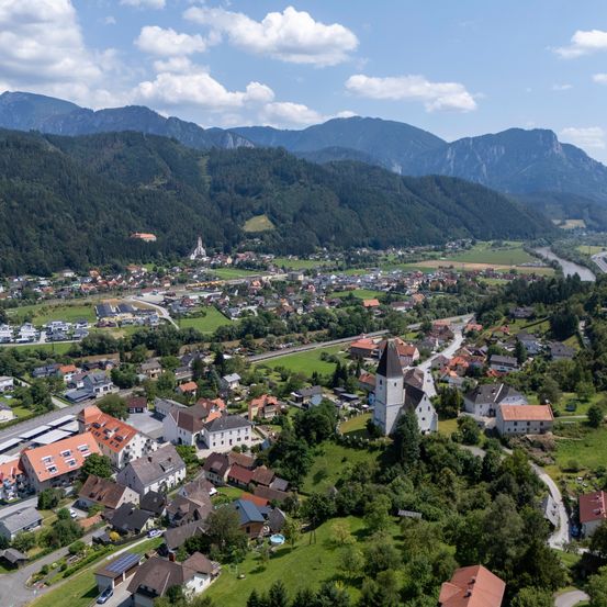 Eine Luftaufnahme eines Dorfes, umgeben von üppiger Vegetation und Bergen, mit einer Kirche und einem Fluss in der Ferne.