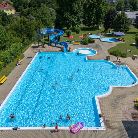 Eine Vogelperspektive auf ein Schwimmbad mit schwimmenden Menschen, Sitzgelegenheiten auf der Terrasse und einer Rutsche mit Schirmen.