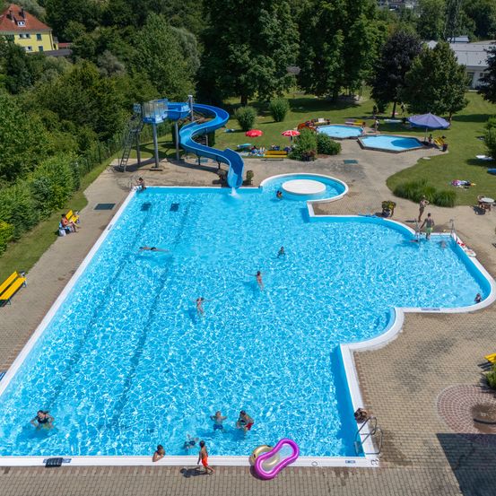 Eine Vogelperspektive auf ein Schwimmbad mit schwimmenden Menschen, Sitzgelegenheiten auf der Terrasse und einer Rutsche mit Schirmen.