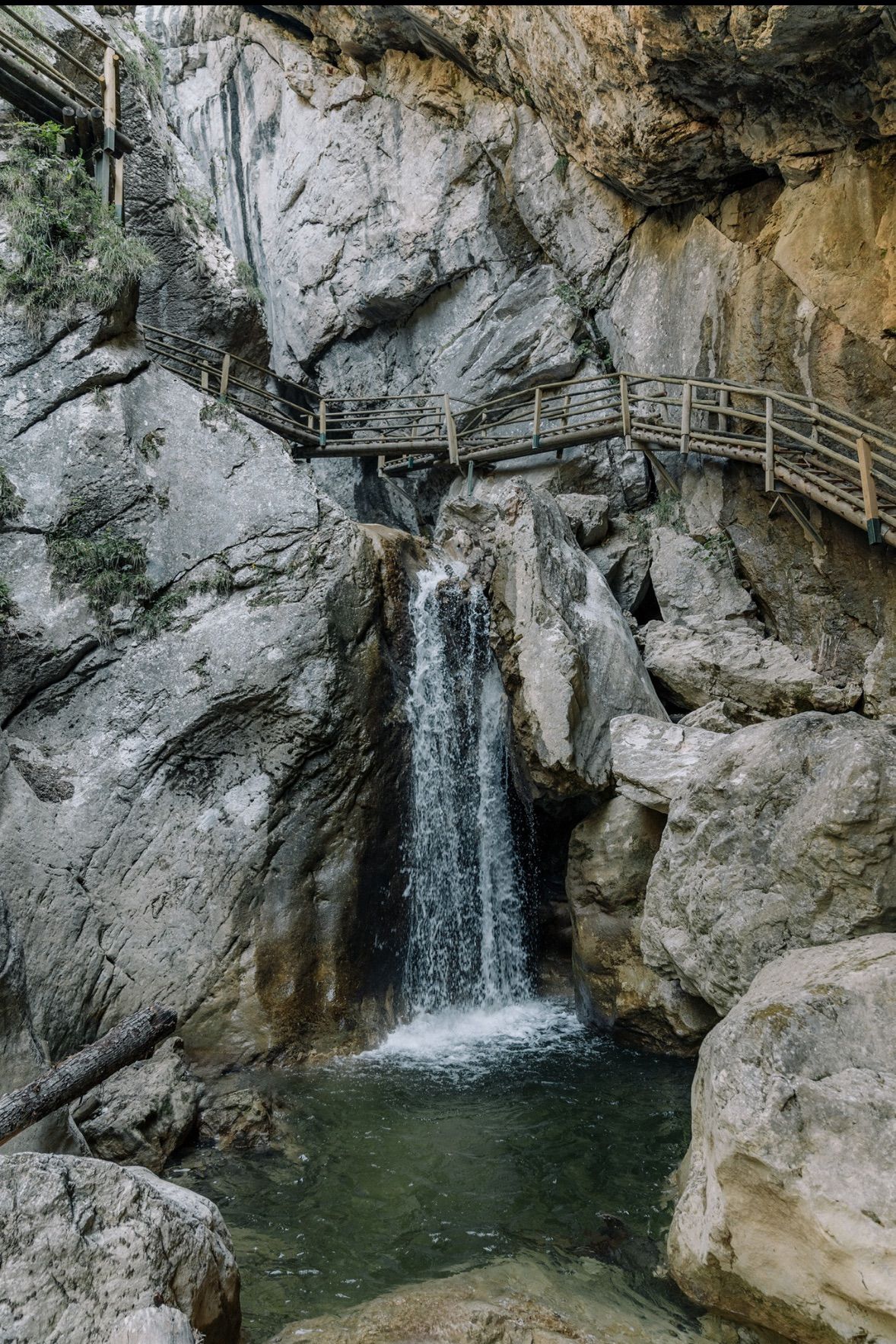 Eine Holzbrücke führt über einen felsigen Abgrund zu einem Wasserfall, der in einen Pool fließt.