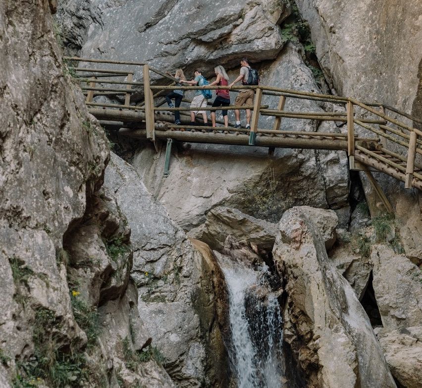 Eine Gruppe von Wanderern überquert eine Holzbrücke über einem Wasserfall in einer felsigen Schlucht.