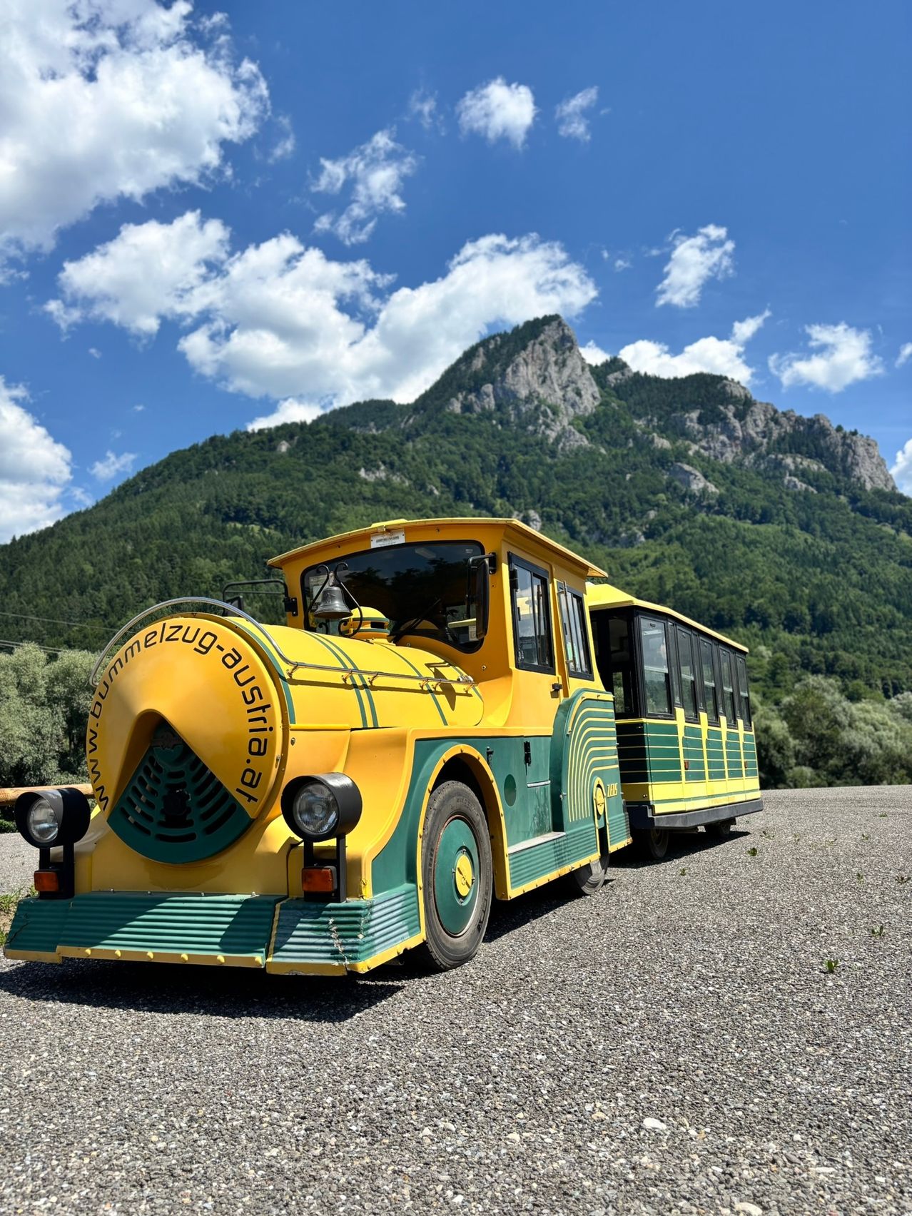 Ein gelber und grüner Zug auf Schotter mit einem Berg und blauem Himmel im Hintergrund.