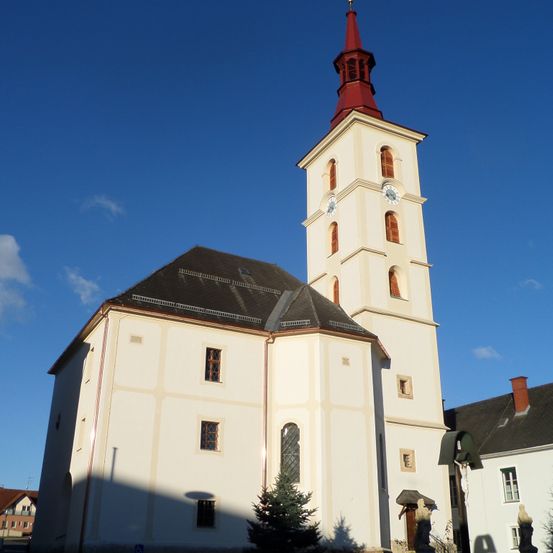 Eine hohe Kirche mit einem roten Turm, einer Uhr auf jeder Seite und mehreren Bogenfenstern. Die Kirche ist weiß mit einem grauen Dach und befindet sich in einer Stadt.