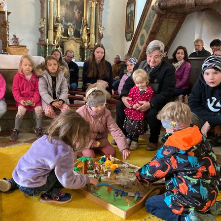 Eine Gruppe von Kindern und Erwachsenen versammelt sich in einer Kirche. Die Kinder spielen mit einem Sandkunsttisch, während die Erwachsenen sitzend zusehen. Der Altar ist mit Blumen geschmückt.