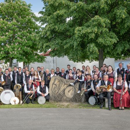 Eine große Gruppe von Musikern, sowohl Männer als auch Frauen, in traditioneller Kleidung, versammelt sich für ein Foto vor einem großen Felsbrocken, mit Bäumen und Gebäuden im Hintergrund.