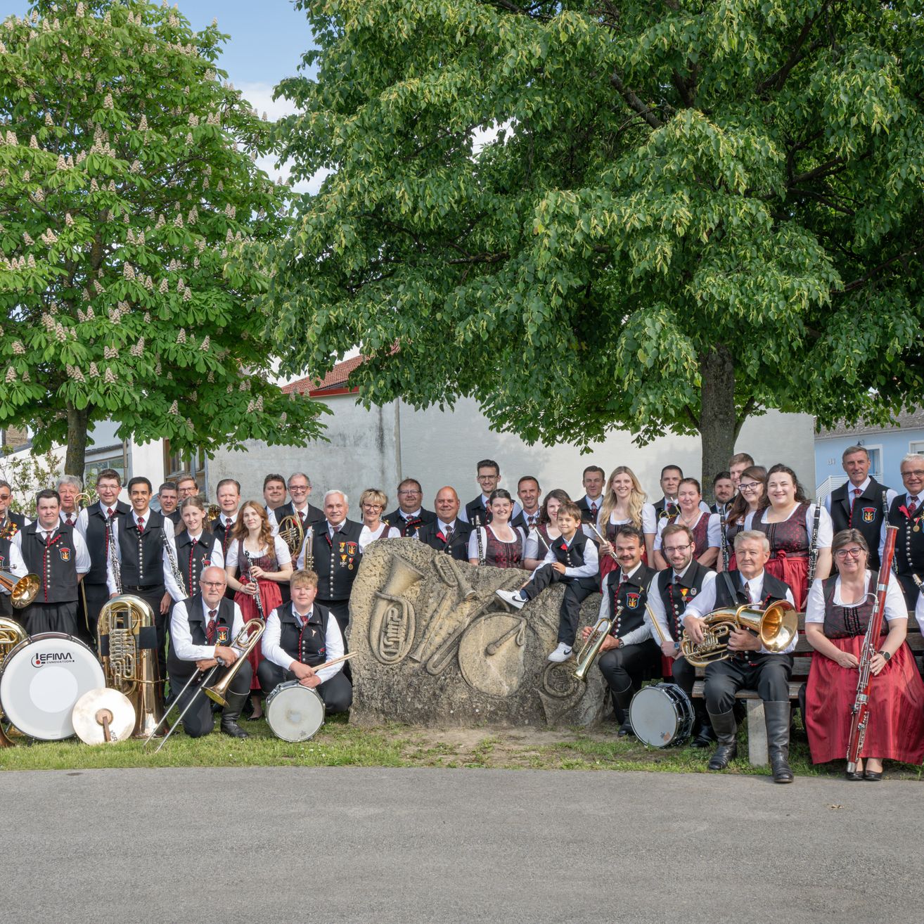 Eine große Gruppe von Musikern, sowohl Männer als auch Frauen, in traditioneller Kleidung, versammelt sich für ein Foto vor einem großen Felsbrocken, mit Bäumen und Gebäuden im Hintergrund.