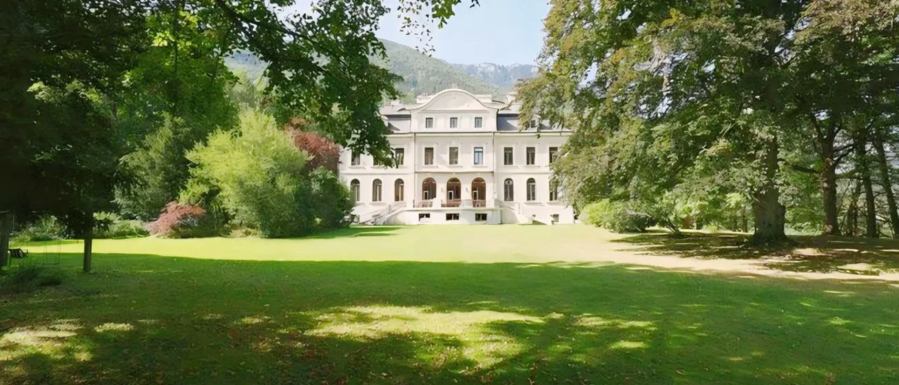 Ein weißes Gebäude steht auf einem üppigen grünen Feld mit Bäumen, die Schatten werfen. Das Gebäude hat viele Fenster und einen großen Eingang. Im Hintergrund sind Berge sichtbar.
