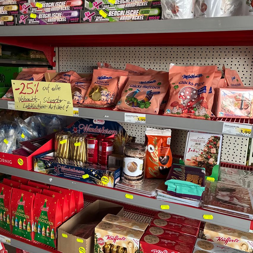 Ein Regal in einem Lebensmittelgeschäft mit verschiedenen Weihnachtsprodukten, darunter Süßigkeiten, Kekse und Geschenkboxen. Ein gelbes Schild wirbt für 25% Rabatt auf alle Weihnachtsartikel.