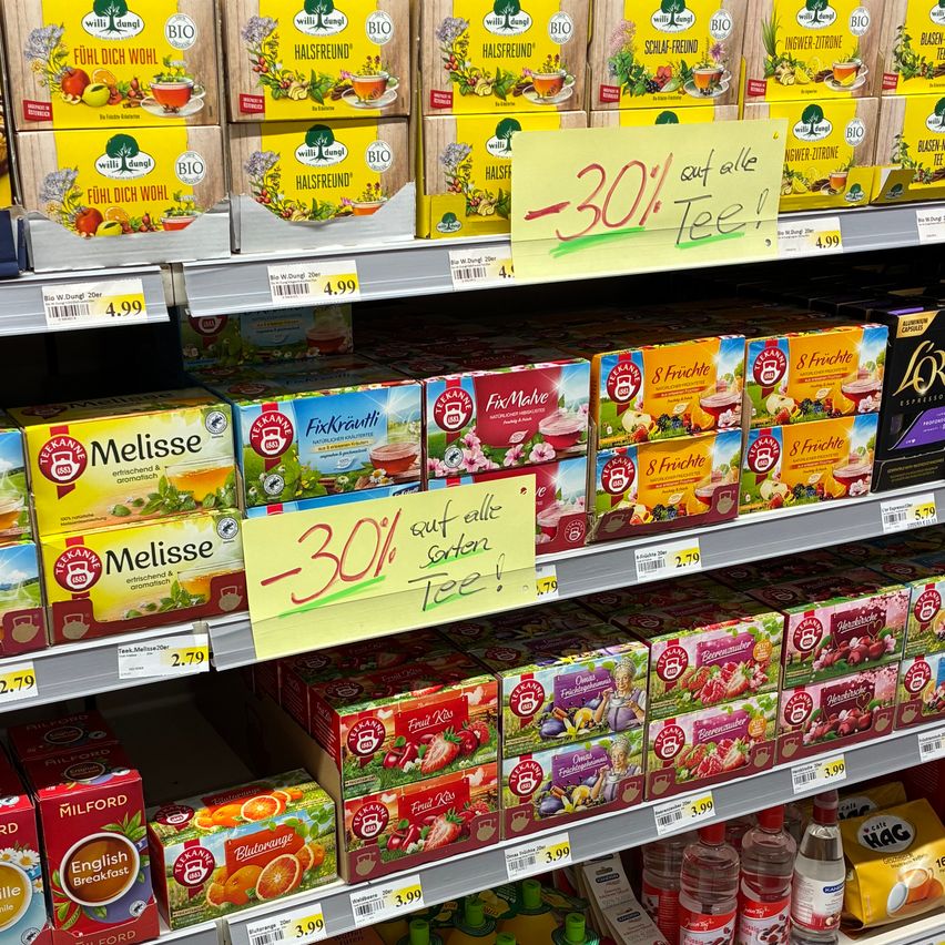 Ein Supermarktregal mit verschiedenen Teeprodukten im Angebot. Ein gelbes Schild weist auf einen 30%igen Rabatt auf alle Tees hin. Schachteln von 'Melisse' und 'FixKräuti' sind prominent ausgestellt. Preise sind auf den Schachteln sichtbar, wobei eine Schachtel einen Preis von 2,79 zeigt.