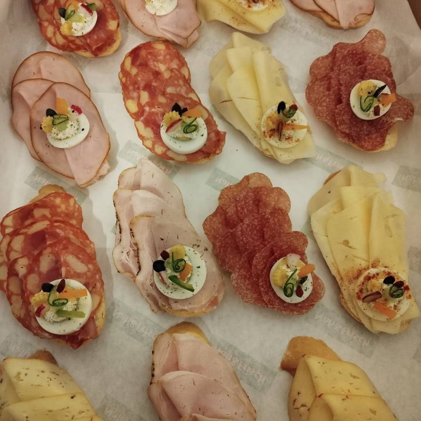 An assortment of sandwiches, each topped with a hard-boiled egg, cheese, and various garnishes. They include slices of ham, salami, and cheese on baguettes.