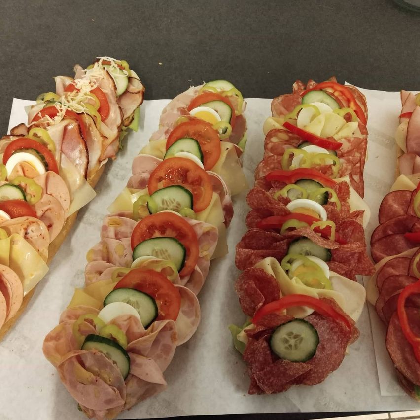 Four sandwiches are on a white paper. Each sandwich is topped with slices of ham, cheese, cucumber, and tomatoes. One sandwich has a red pepper slice on top.