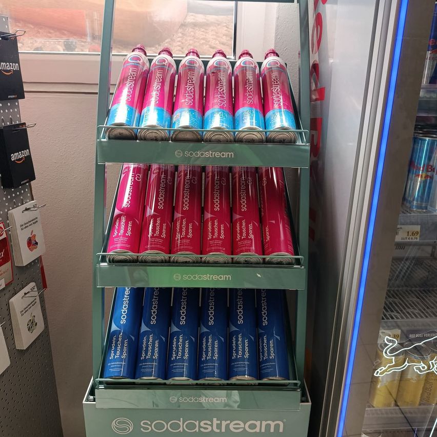 Eine Display von SodaStream Dosen in verschiedenen Geschmacksrichtungen und Farben ist in einem Laden zu sehen. Die Dosen sind ordentlich auf grünen Regalen an einer Wand mit Amazon-Logos gestapelt.