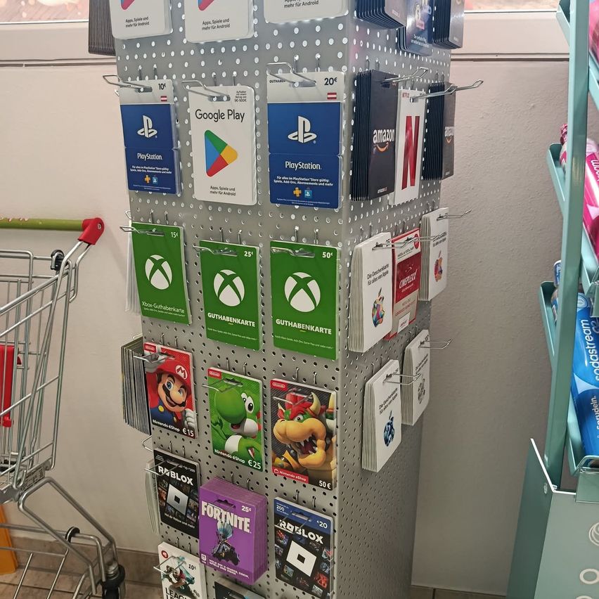 Ein Display mit verschiedenen Videospiel-Geschenkkarten, darunter PlayStation, Google Play, Xbox, Amazon und Fortnite. Die Karten sind an einem Metall-Stiftleiste befestigt. Ein Einkaufswagen steht neben dem Display.