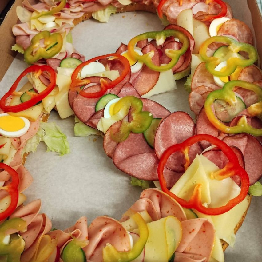 Ein runder Sandwich in einer Box mit Schinken, Käse, geschnittenen Gurken und roten und grünen Paprikaschoten.