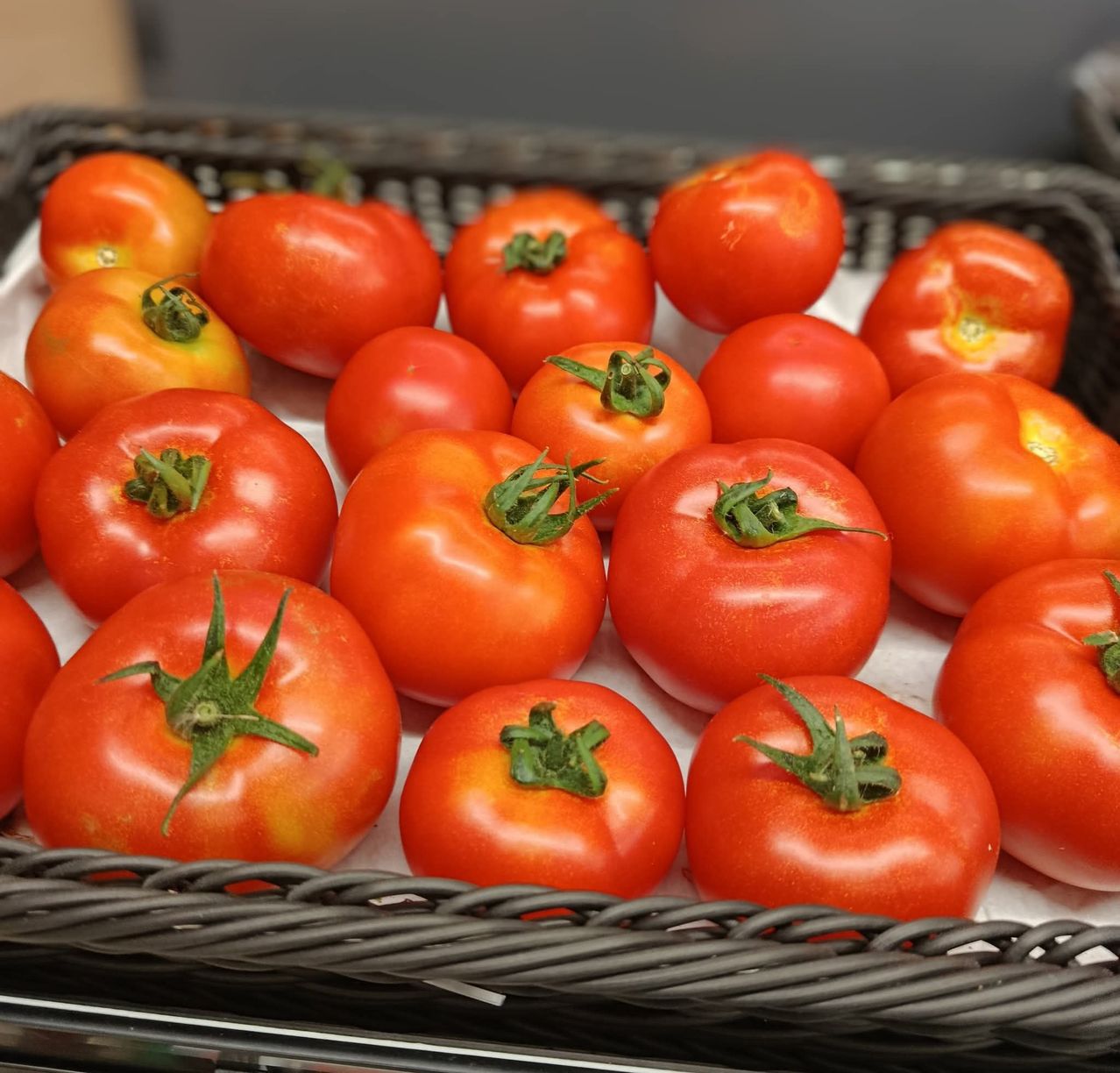 Ein Korb voller roter Tomaten mit grünen Stielen, ordentlich auf einer weißen Oberfläche angeordnet. Jede Tomate ist rund und leuchtend.
