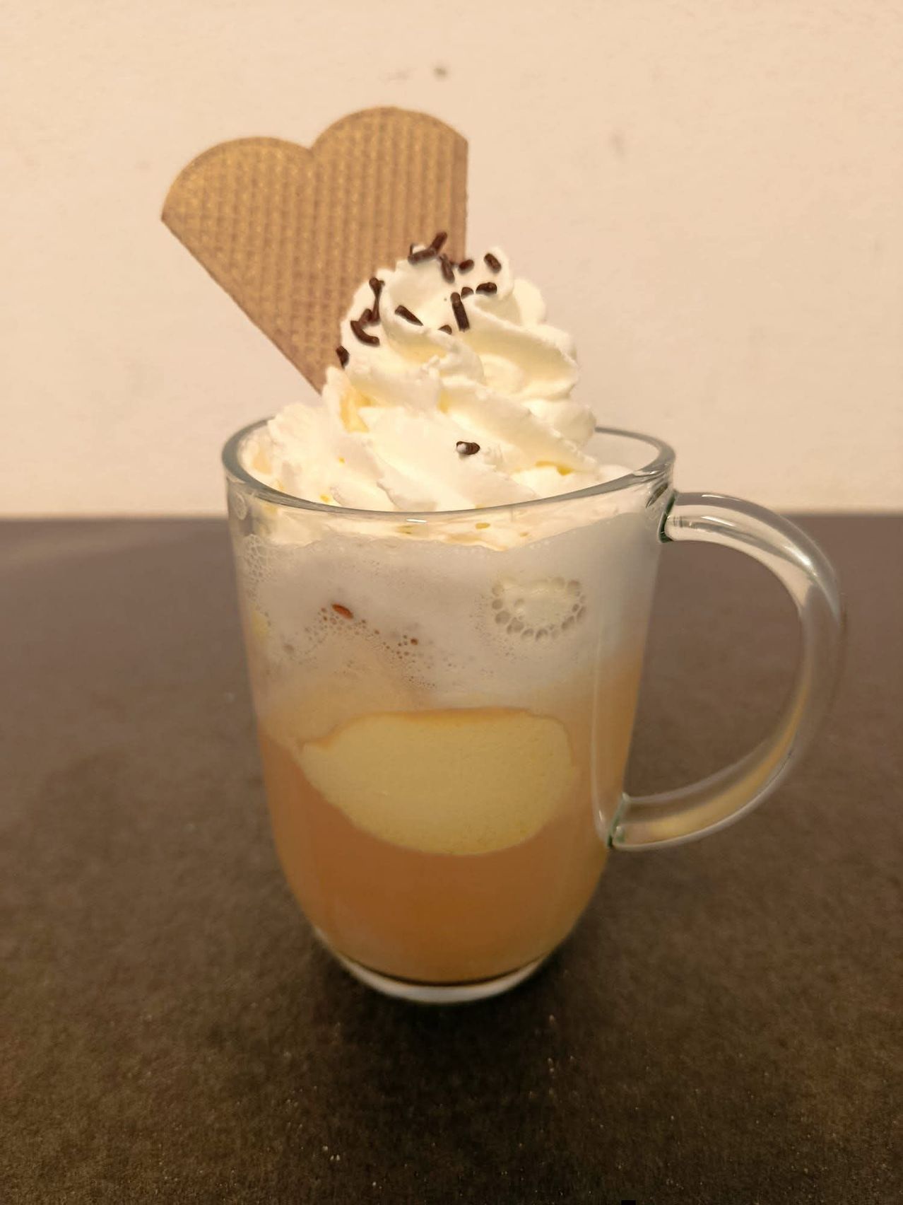 Auf einem Tisch steht ein Glas mit Schlagsahne, Schokostückchen und einem Waffelkegel darauf. Das Getränk darin ist cremig und hat ein Apfelstück.