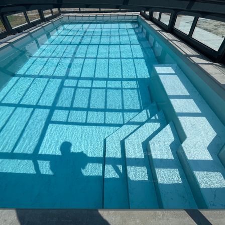 Ein klares, rechteckiges Schwimmbecken mit Glaswänden und einem gefliesten Boden. Der Schatten einer Person wird von oben auf das Becken geworfen.