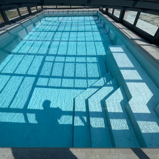 Ein klares, rechteckiges Schwimmbecken mit Glaswänden und einem gefliesten Boden. Der Schatten einer Person wird von oben auf das Becken geworfen.