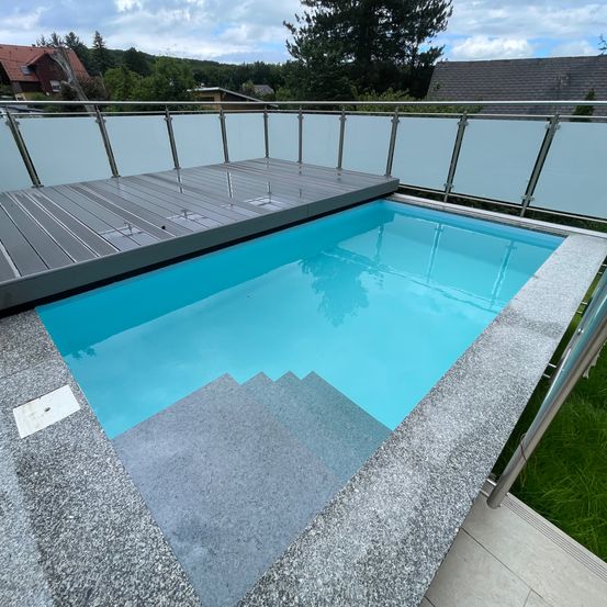 Ein Freibad mit einer blauen Wasseroberfläche, umgeben von einem grauen Steinrand und einer Holzterrasse. Ein Metallgeländer ist an der Holzterrasse befestigt.