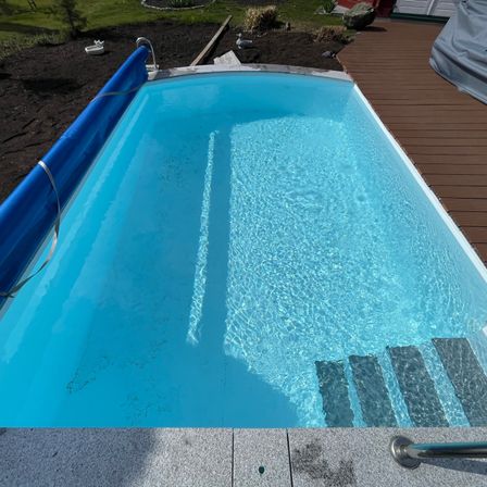 Ein Außenpool mit blauer Umrandung und klarem Wasser. Er hat eine Holzterrasse mit Stufen, die in den Pool führen.