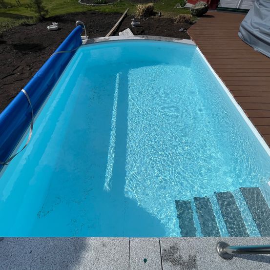 Ein Außenpool mit blauer Umrandung und klarem Wasser. Er hat eine Holzterrasse mit Stufen, die in den Pool führen.