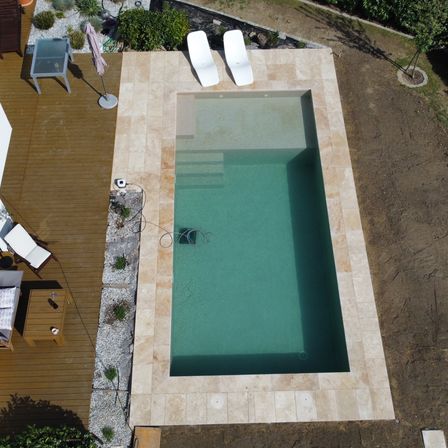 Eine Vogelperspektive eines modernen, rechteckigen Pools, umgeben von einer Steinumrandung, mit zwei weißen Liegestühlen und einem kleinen Tisch auf einer Holzterrasse.