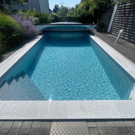 Ein rechteckiger Swimmingpool mit klarem blauem Wasser, umgeben von einem grauen Betonrand. Ein transparenter Deckel befindet sich über dem Pool, mit Stufen auf einer Seite. Der Pool befindet sich in einem Garten mit Pflanzen und einem Holzzaun.
