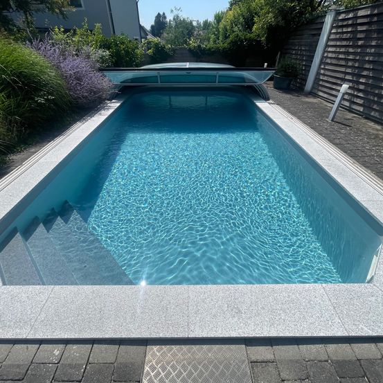 Ein rechteckiger Swimmingpool mit klarem blauem Wasser, umgeben von einem grauen Betonrand. Ein transparenter Deckel befindet sich über dem Pool, mit Stufen auf einer Seite. Der Pool befindet sich in einem Garten mit Pflanzen und einem Holzzaun.