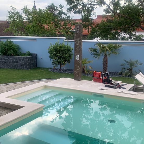 Ein Freibad mit Betondeck und Duschkopf. Der Pool ist von einem Garten mit verschiedenen Pflanzen umgeben.