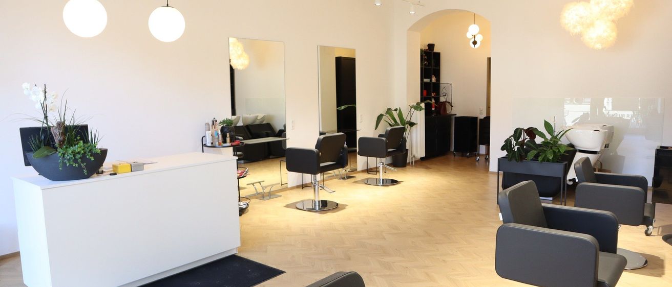 Ein Salon mit mehreren schwarzen Stühlen, Spiegeln und einer weißen Theke. Der Raum hat Holzböden und weiße Wände. Es gibt Regale mit Pflanzen und Dekorationsartikeln.