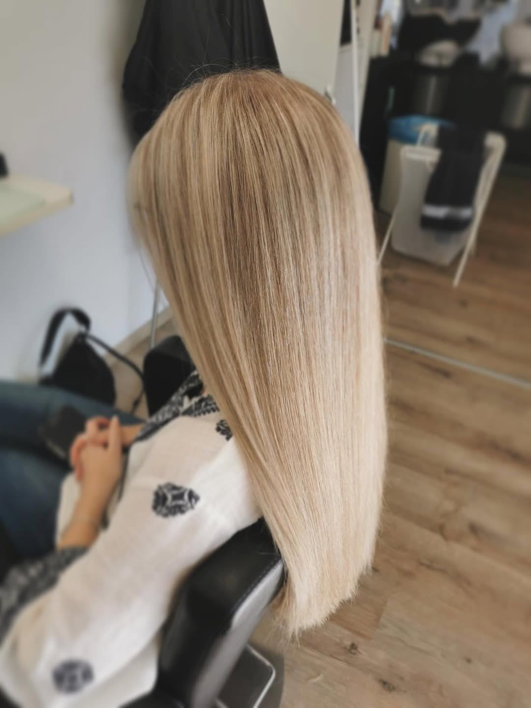 Eine Frau mit langen blonden Haaren sitzt in einem Salonstuhl und schaut auf etwas auf dem Boden. Hinter ihr befindet sich ein weißer Tisch und ein Plastikbehälter.
