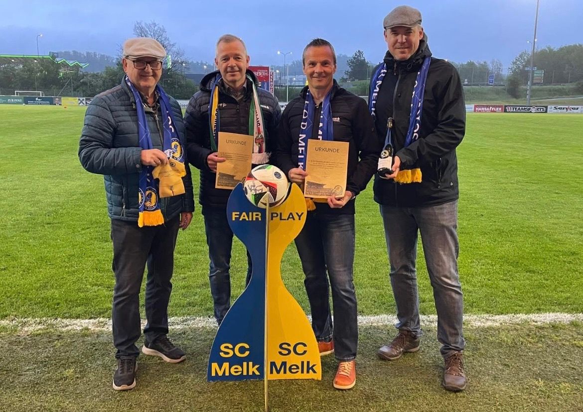 Vier Männer stehen auf einem Fußballplatz, lächeln und halten Urkunden und einen Fußball. Der Mann auf der rechten Seite hält eine Flasche Wein. Hinter ihnen steht ein Banner, das 'Fair Play' und 'SC Melk' besagt.