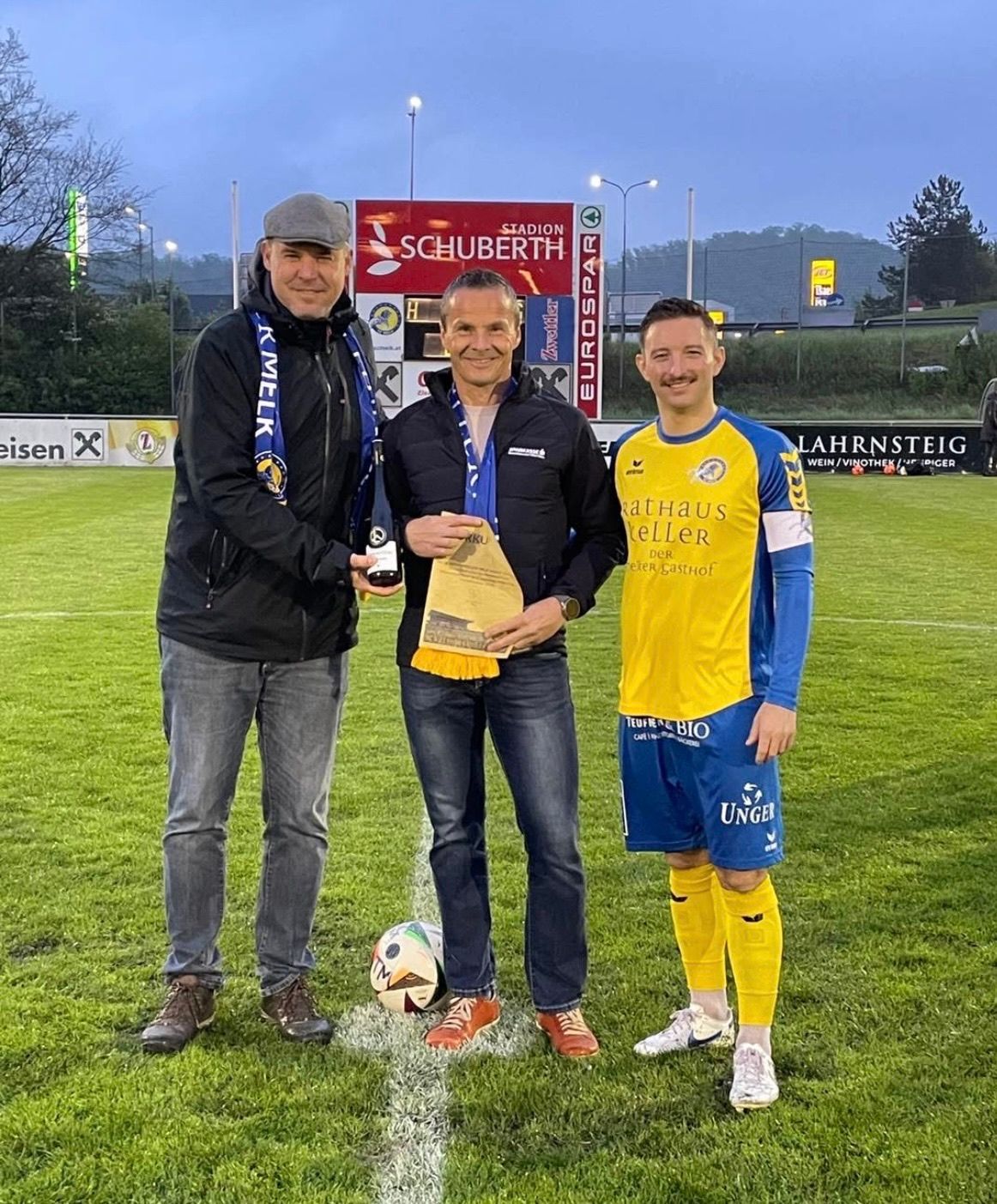 Drei Männer stehen auf einem Fußballfeld, zwei von ihnen tragen Trikots. Der Mann auf der linken Seite hält eine Flasche, und der Mann in der Mitte hält ein Stück Papier. Der Mann auf der rechten Seite lächelt und trägt ein gelbes Trikot.