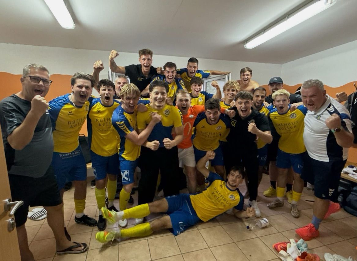 A soccer team in blue and yellow uniforms celebrates in a locker room, with one player lying on the floor and others cheering.