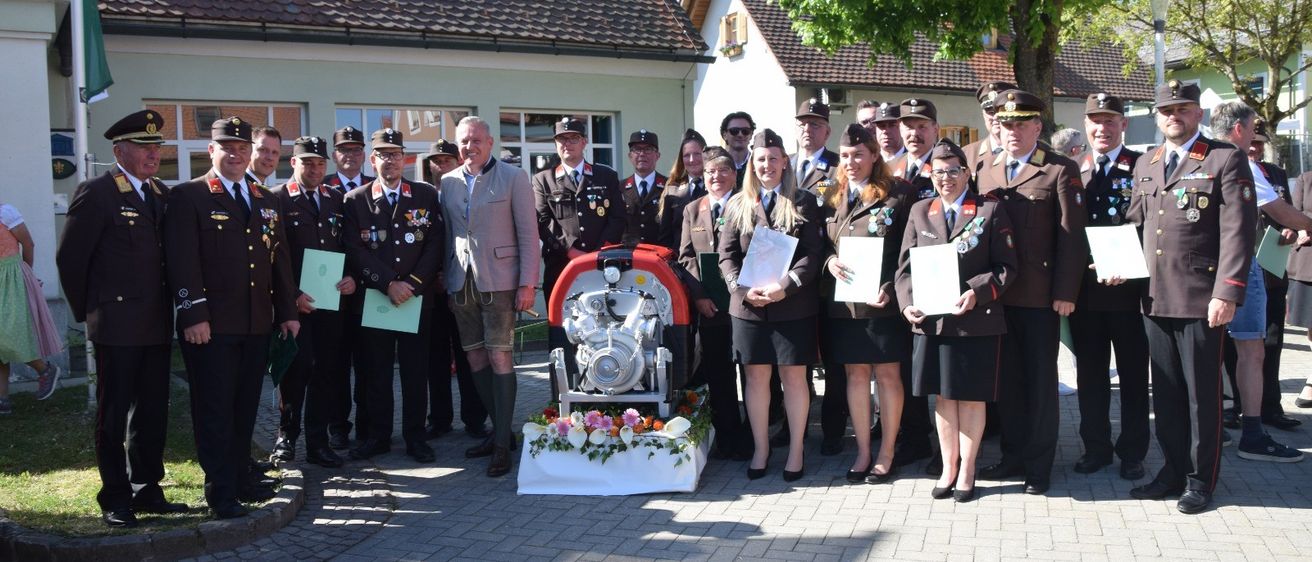 Eine Gruppe von Menschen in Uniformen, einschließlich Männer und Frauen, stehen vor einem Haus mit einer Maschine und Blumen. Sie halten Urkunden und schauen zur Kamera.
