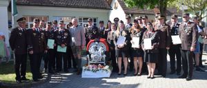 Eine Gruppe von Menschen in Uniformen, einschließlich Männer und Frauen, stehen vor einem Haus mit einer Maschine und Blumen. Sie halten Urkunden und schauen zur Kamera.