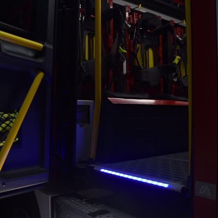 Innenansicht eines Feuerwehrwagens mit offenen Türen. Gelbe Handläufe und ein blauer LED-Streifen beleuchten das Innere. Feuerwehrausrüstung ist im Hintergrund ordentlich angeordnet.