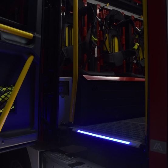 Innenansicht eines Feuerwehrwagens mit offenen Türen. Gelbe Handläufe und ein blauer LED-Streifen beleuchten das Innere. Feuerwehrausrüstung ist im Hintergrund ordentlich angeordnet.