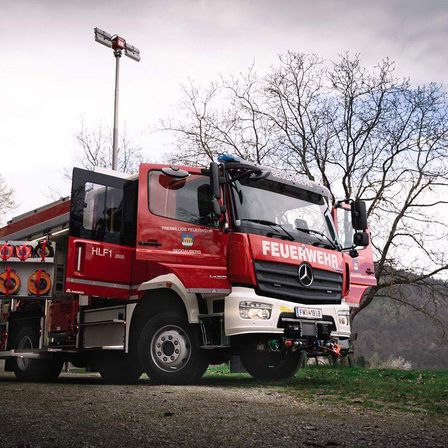 Ein roter Mercedes-Benz Feuerwehrwagen, gekennzeichnet mit HLF1 2000, steht im Freien mit geöffneter Tür. Er ist mit verschiedenen Geräten ausgestattet und hat eine angebrachte Leuchte auf einem Mast dahinter.