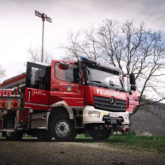 Ein roter Mercedes-Benz Feuerwehrwagen, gekennzeichnet mit HLF1 2000, steht im Freien mit geöffneter Tür. Er ist mit verschiedenen Geräten ausgestattet und hat eine angebrachte Leuchte auf einem Mast dahinter.