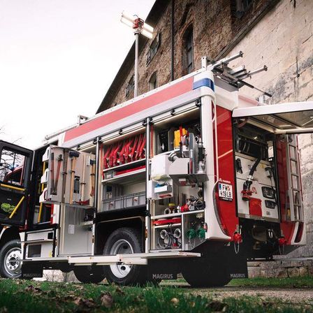 Ein roter und weißer Feuerwehrwagen mit offenen Hecktüren steht vor einem Backsteingebäude. Der Wagen ist mit verschiedenen Werkzeugen und Ausrüstungsgegenständen im Inneren ausgestattet.