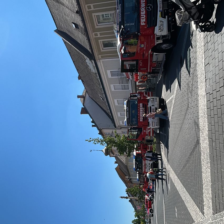 Feuerwehrfahrzeuge sind auf der Straße vor Gebäuden geparkt. Eine Person steht in der Nähe der Fahrzeuge. Der Himmel ist klar und blau.