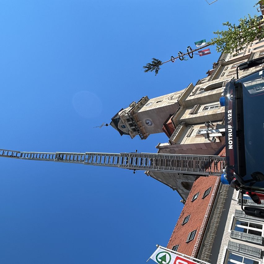 Ein Feuerwehrwagen mit ausgefahrener Leiter zu einem Gebäude mit Flaggen und einem Uhrturm unter einem klaren blauen Himmel.