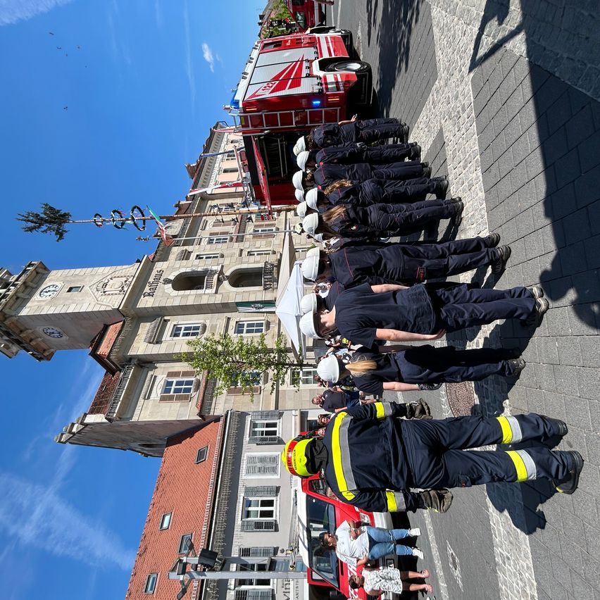 Eine Gruppe von Feuerwehrleuten steht in Formation vor einem Gebäude mit Glockenturm. Die Sonne wirft Schatten auf den Bürgersteig, und ein roter Feuerwehrwagen ist in der Nähe geparkt.