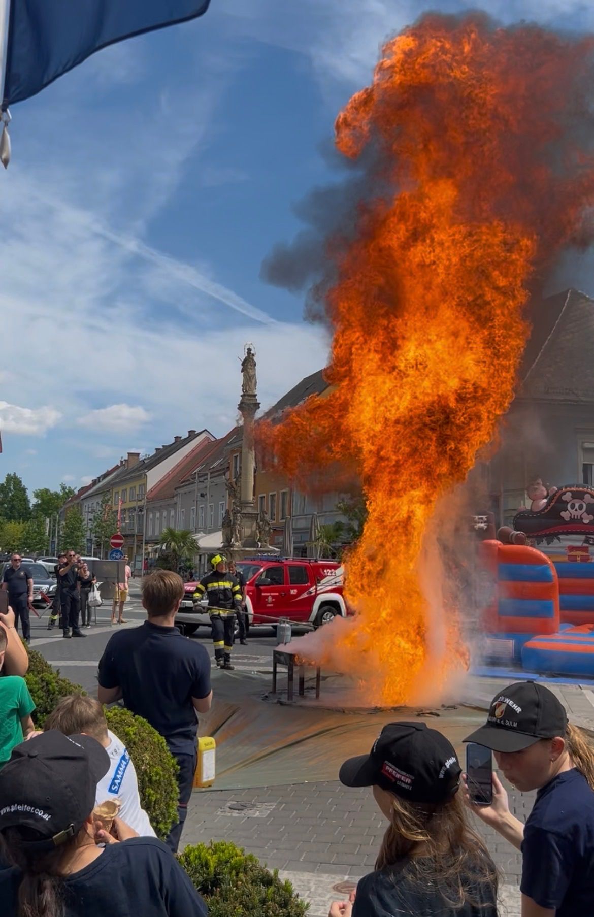 Auf einer Straße findet eine Feuervorführung statt, mit Zuschauern, Feuerwehrleuten und einer großen Flamme in der Mitte. Im Hintergrund sind Gebäude und Autos zu sehen.