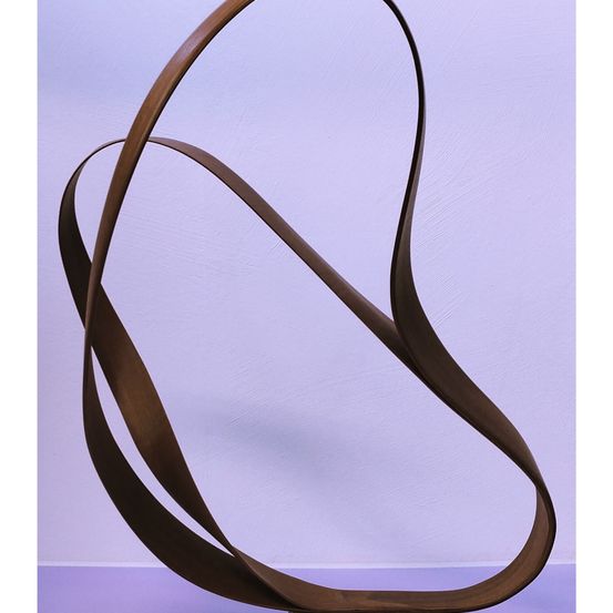 Eine bronzene Skulptur mit kurvigem, abstraktem Design steht vor einem lila Hintergrund. Die Skulptur besteht aus mehreren sich überschneidenden Bögen, die ein dynamisches und fließendes Erscheinungsbild erzeugen.