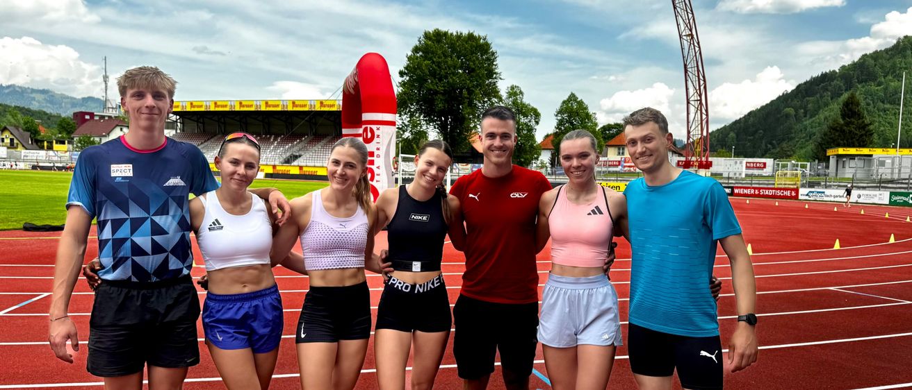 Fünf Athleten stehen auf einer Laufbahn und lächeln für ein Foto. Ein Mann mit einem roten Shirt steht in der Mitte, während vier Frauen um ihn herum stehen und verschiedene farbige Oberteile und Shorts tragen.
