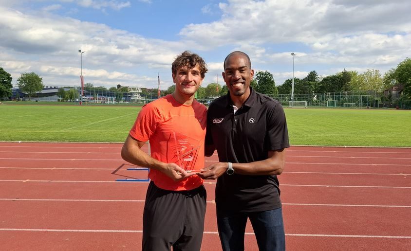 Zwei Männer stehen auf einer Laufbahn, einer hält eine Trophäe. Der Mann in Orange lächelt und der Mann in Schwarz trägt eine Uhr. Sie sind umgeben von einem Rasenfeld und einem Zaun.