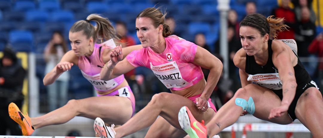 Drei Athletinnen konkurrieren in einem Hürdenrennen. Die führende Athletin trägt ein rosa Trikot mit dem Namen 'TRAMETZ'. Zuschauer beobachten von den Tribünen.