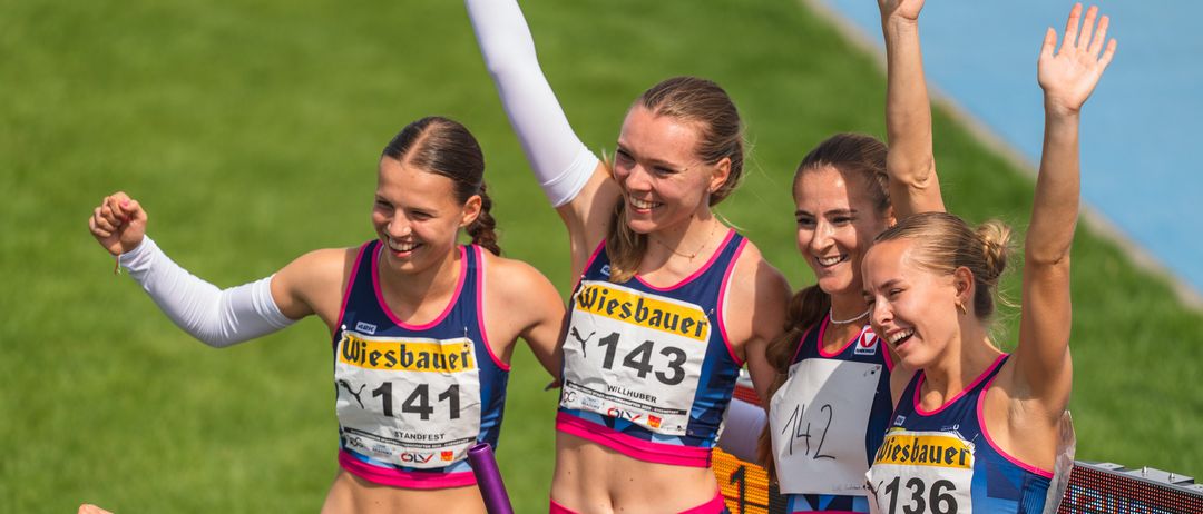 Mehrere Läuferinnen in blauen und rosa Uniformen mit Nummern feiern auf der Laufbahn. Ein Mann vor ihnen mit geballter Faust lächelt.